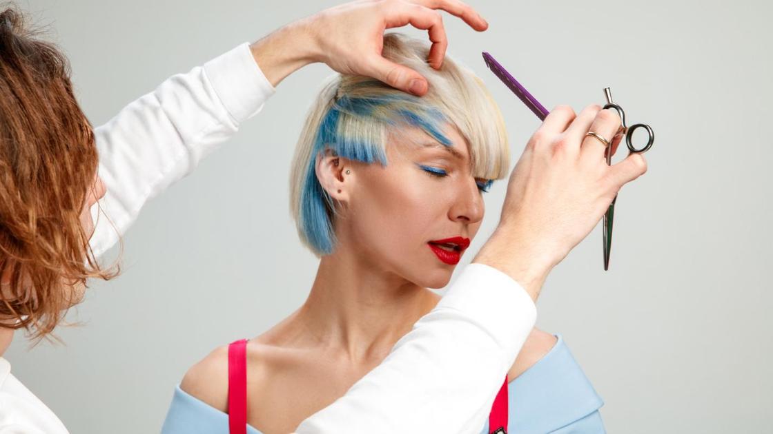 Парикмахер делает стрижку на белокурых волосах со скрытым окрашиванием в голубом оттенке