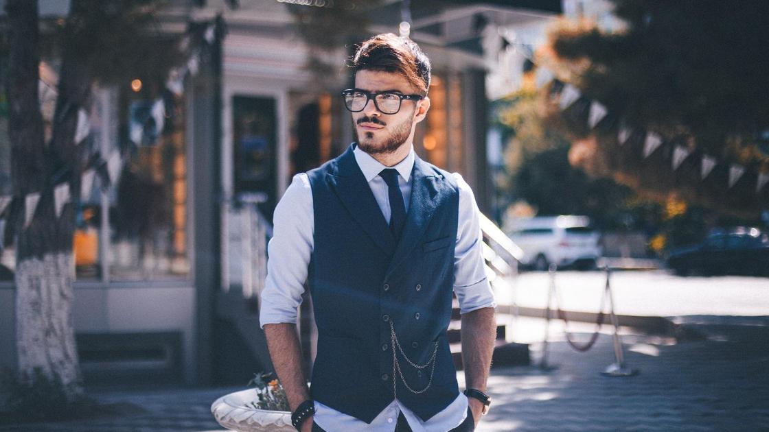 Мужчина в жилетке, галстуке и очках стоит на улице