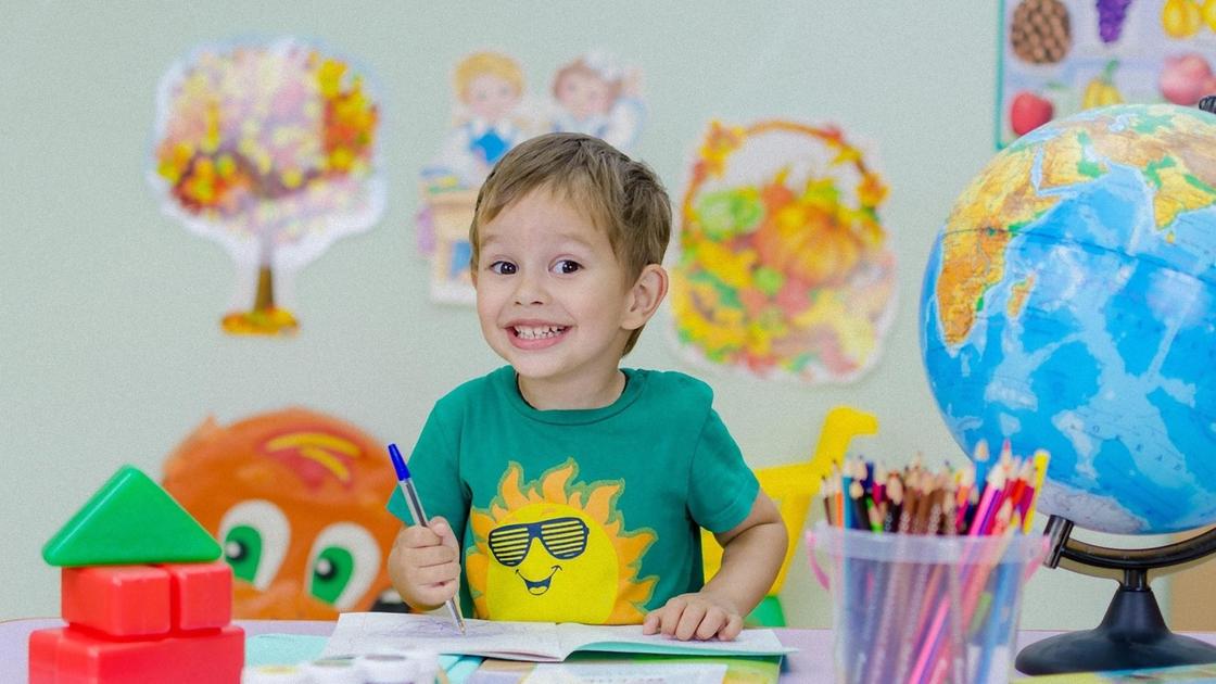 Улыбающийся мальчик в зеленой футболке с принтом в виде солнца стоит возле стола и держит в руках ручку. На столе стоит коробка с карандашами, глобус, кубики и лежит раскрытая тетрадь