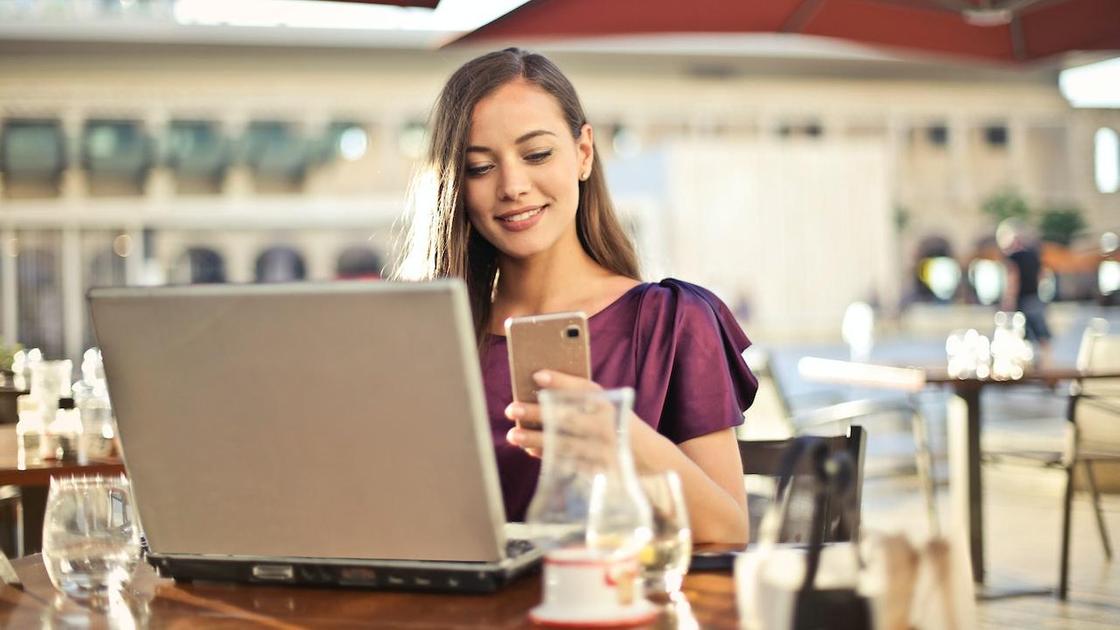 Девушка с телефоном в руках работает за ноутбуком в кафе