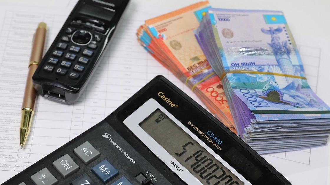 Пачка денег лежит на графике рядом с калькулятором, телефоном и ручкой