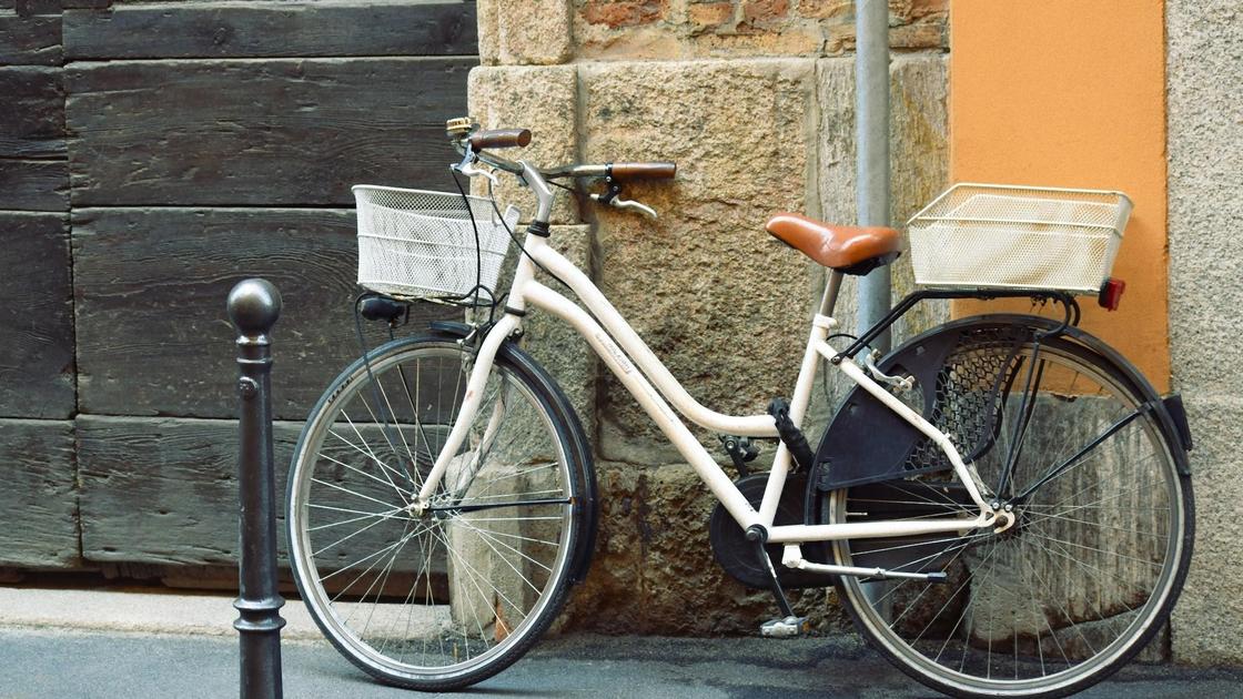 Под стеной дома стоит женский велосипед с рамой белого цвета, корзиной спереди и багажником сзади