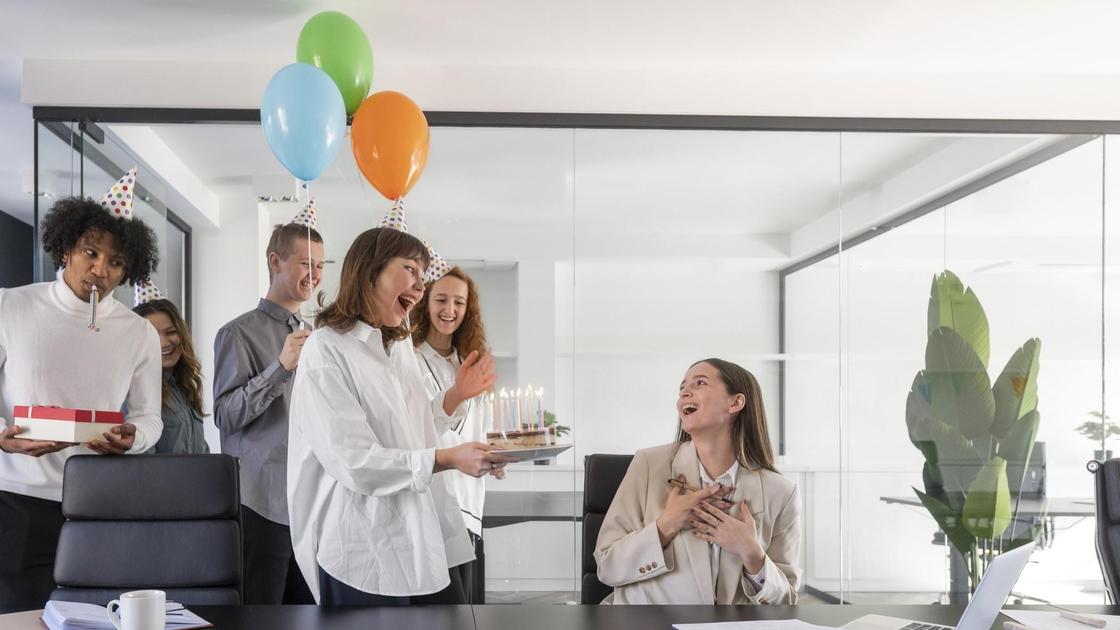 Парни и девушки поздраляют коллегу по работу с днем рождения. Именинница сидит и улыбается, а ей вручают торт со свечами, подарок и шарики
