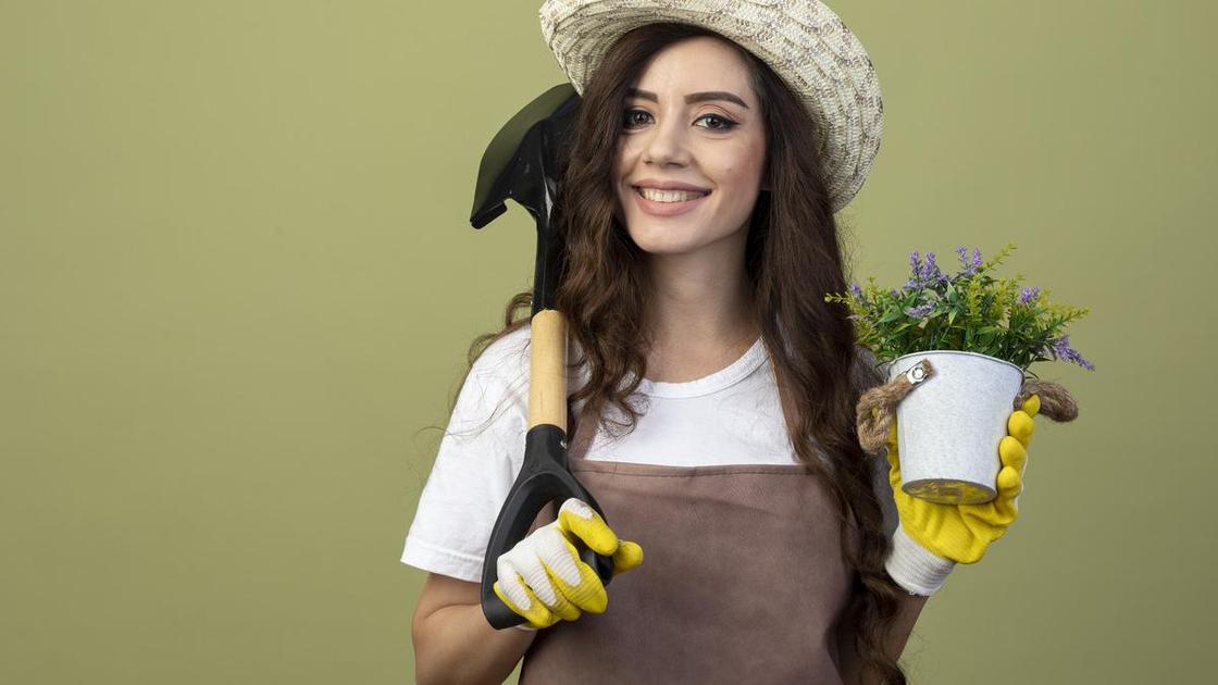Девушка в шляпе и фартухе с садовой лопатой на плече и цветущими растениями в горшке