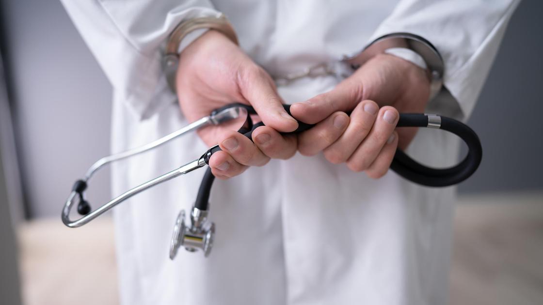 Человек в медицинском халате в наручниках держит за спиной стетоскоп