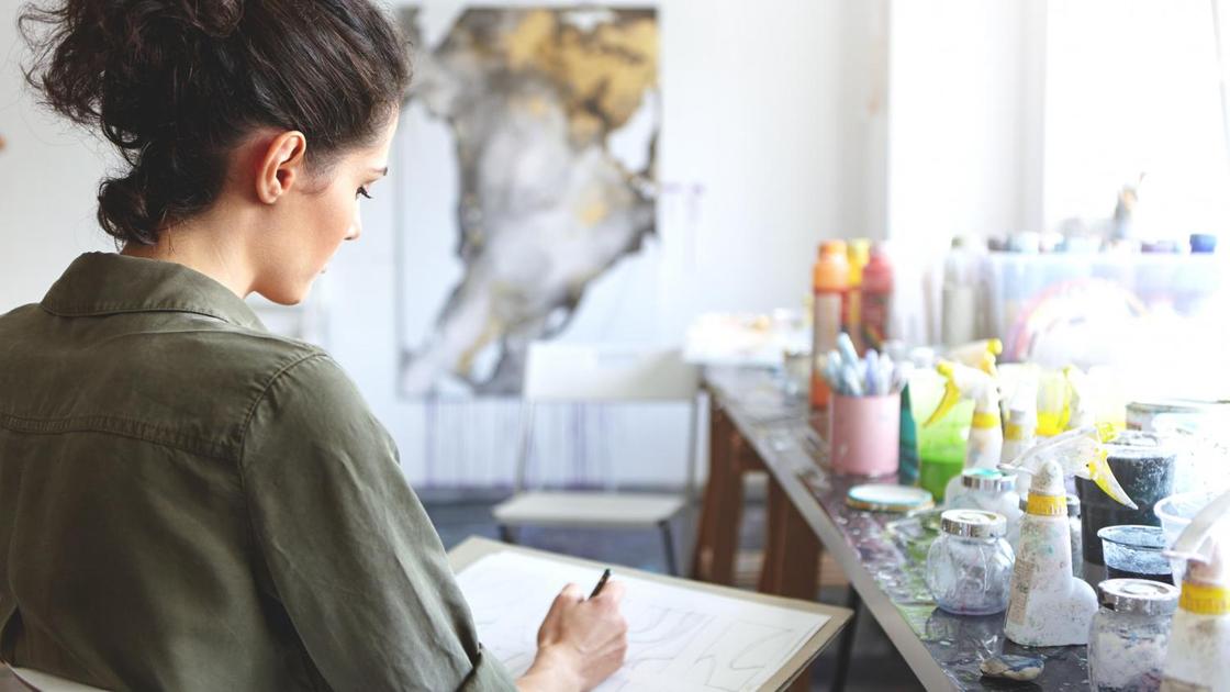 Девушка рисует простым карандашом на бумаге сидя у стола с красками
