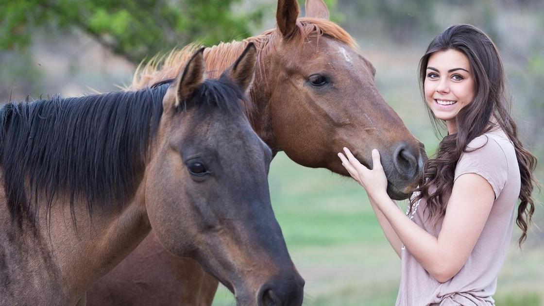 Девушка с длинными волосами стоит возле двух лошадей. Одну из них она держит за морду