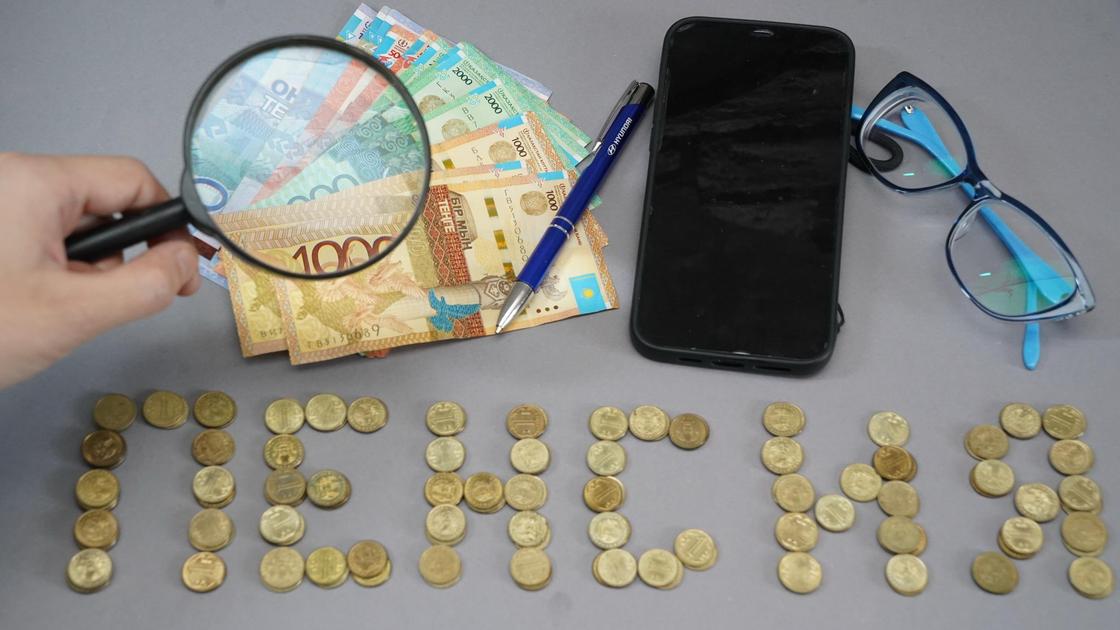 Монеты и купюры тенге лежат рядом со смартфоном и очками