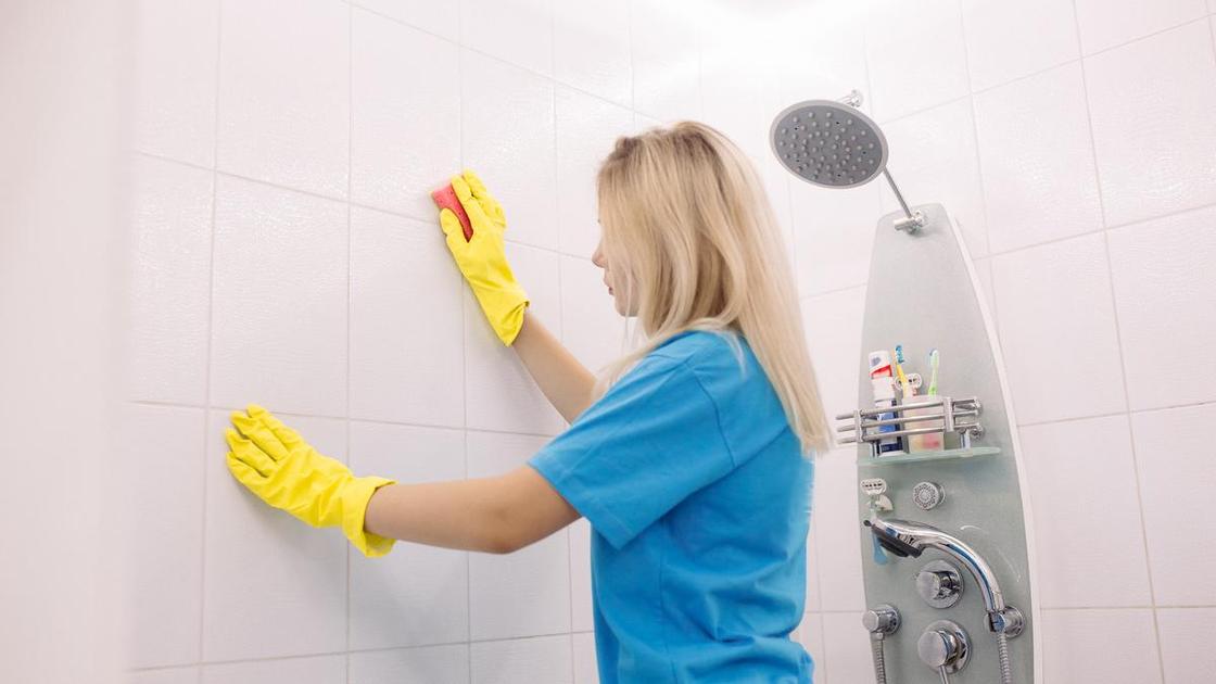 Девушка в синем костюме и желттых резиновых перчаткает моет плитку на стене в ванной
