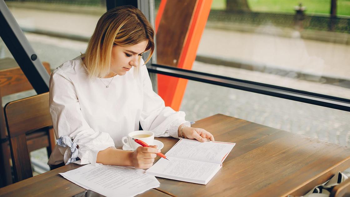 Девушка с ручкой в руках сидит за столом и читает блокнот
