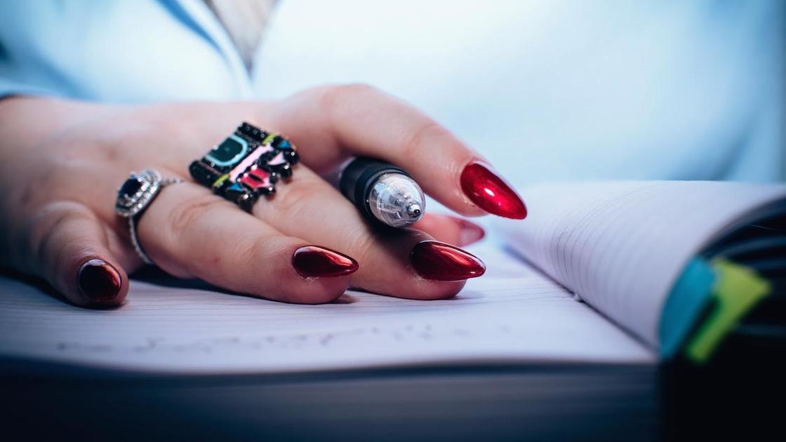 Женская рука с красивым зеркальным маникюром в красном цвете держит ручку