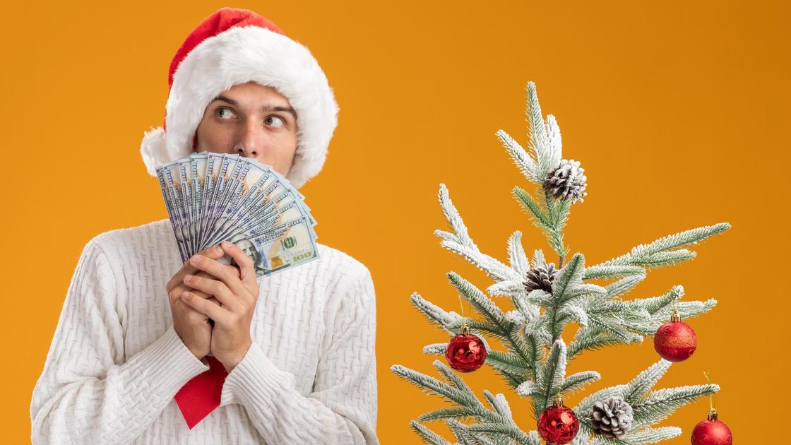 Парень в новогодней шапке прячет лицо за веером из денег