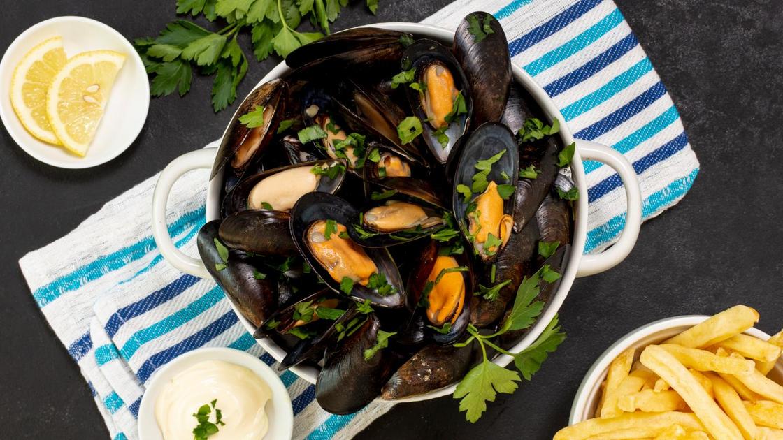 В сковороде мидии с зеленью в ракушках. Рядом пиалы с дольками лимонаи соусами