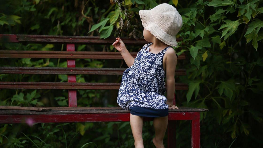 Маленькая девочка в платье и шляпке сидит на скамейке