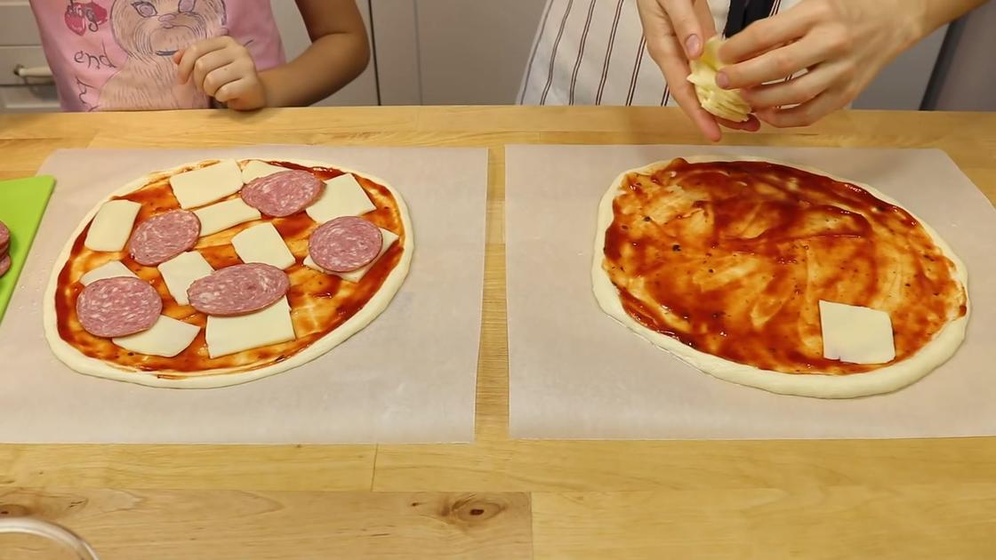 На столе две заготовки для пиццы. Одна намазана томатным соусом, вторая наполнена салями, соусом и сыром