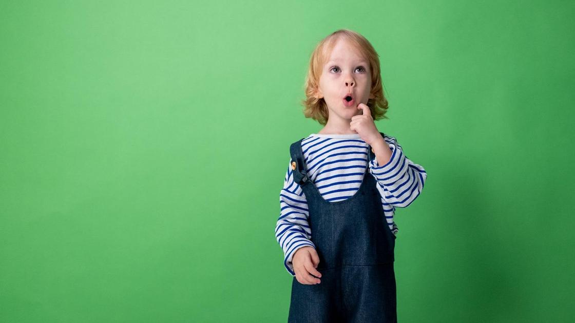 Удивленный ребенок в полосатой кофте и джинсовом комбинезоне на зеленом фоне
