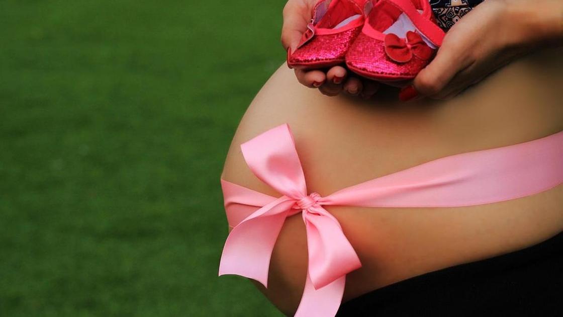 Беременная женщина с оголенным животом перевязанным розовой лентой держит розовые пинетки