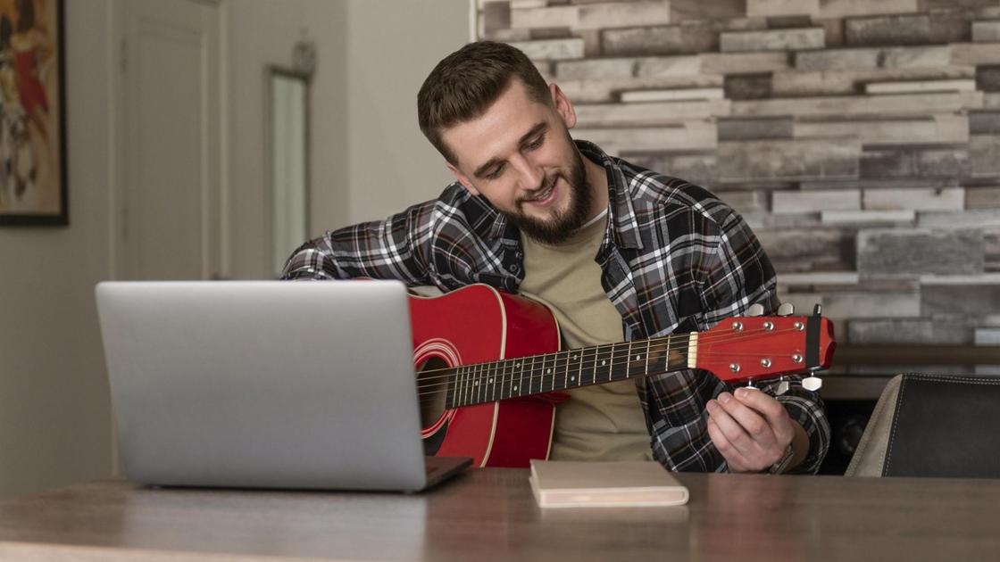 Бородатый мужчина сидит перед открытым ноутбуком и настраивает гитару
