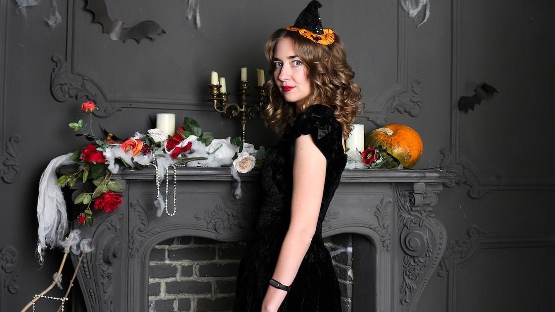 Девушка с костюме ведьмы стоит у камина с атрибутикой Хэллоуина