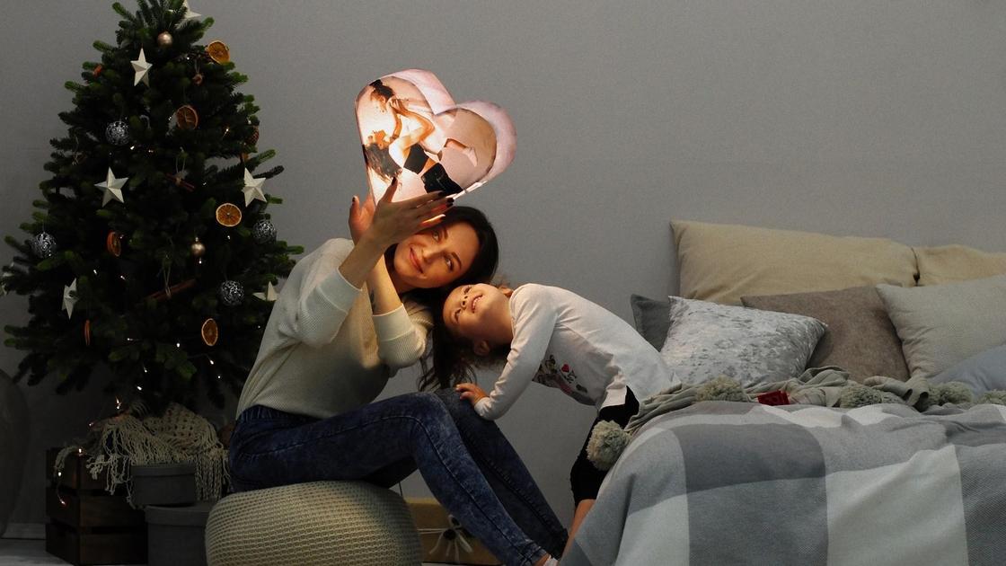 Дочка с мамой сидят возле новогодней елки и рассматривают большое сердце с подсветкой и фотографией. Мама сидит на пуфике, а дочка на кровати с подушками и пледом
