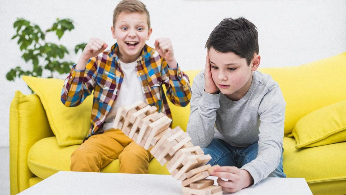 Два мальчика сидят и играют в джангу. Один мальчик вытягивает деревянный брусок и башня падает