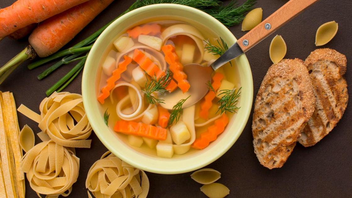 На столе тарелка с куриным супом и лапшой. Рядом гренки, морковь и высушенная лапша