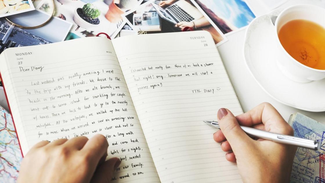 Женщина делает записи ручкой в открытом дневнике. Рядом стоит на блюдце белая чашка с чаем