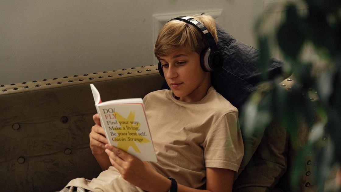 Подросток в наушниках читает книгу, лежа на диване