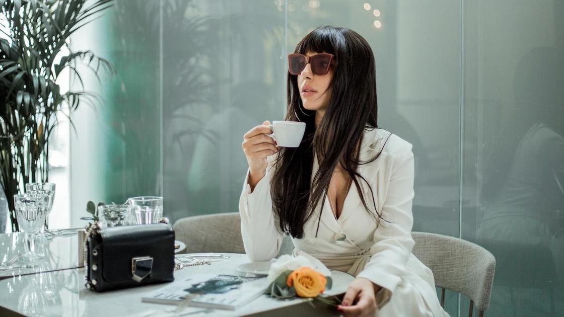 Женщина в белом пьет кофе, сидя за столиком