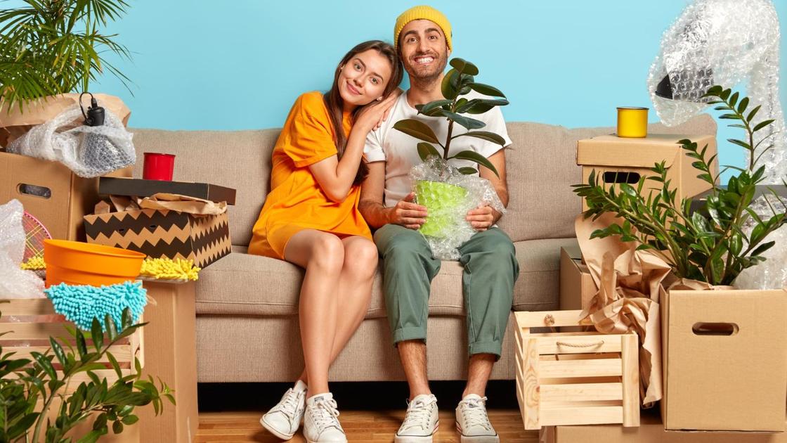 Молодая пара на диване: парень держит в руках горшок с фикусом. Вокруг зеленые растения и ящики