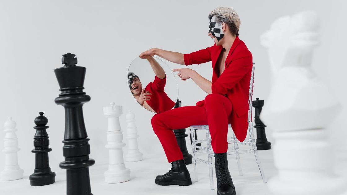Молодой человек в красном костюме смотрит в зеркало рядом с шахматными фигурами