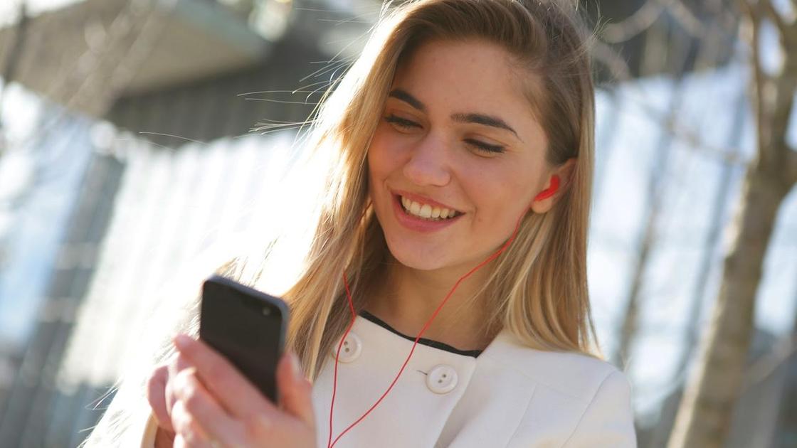 Девушка в наушниках улыбается, глядя на телефон Девушка в наушниках улыбается, глядя на телефон