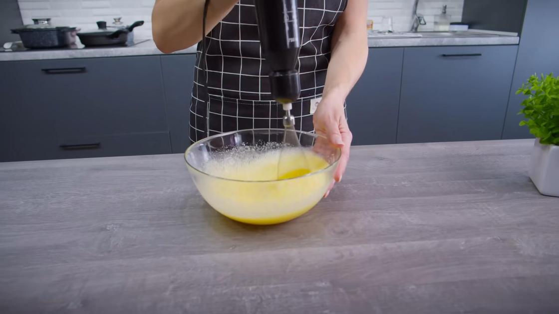 Девушка в фартуке блендером взбивает яично-сахарную массу в стеклянной миске