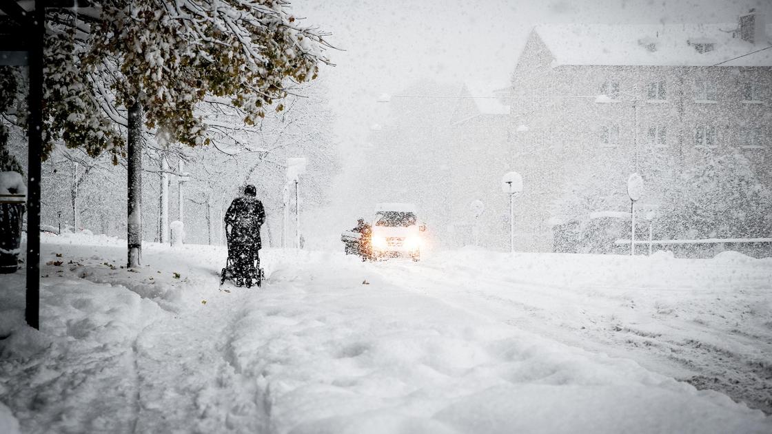 Человек идет по улице во время снегопада на фоне деревьев, зданий и автомобиля