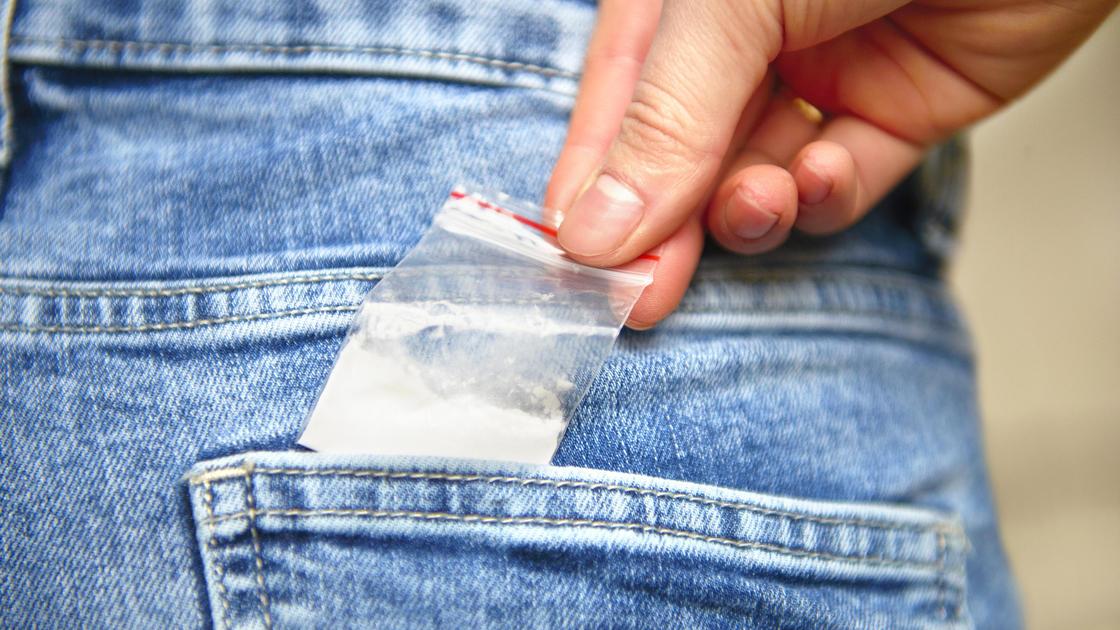 Человек достает пластиковый пакетик с белым порошком из заднего кармана на джинсах
