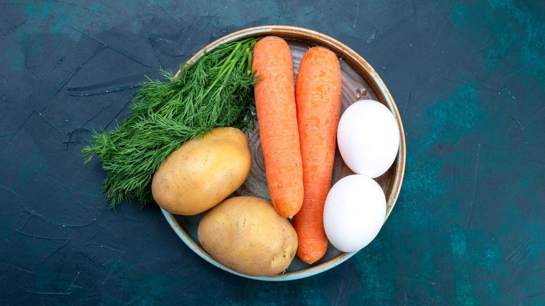 На тарелке сырые картофель, морковь и яйца с пучком зелени