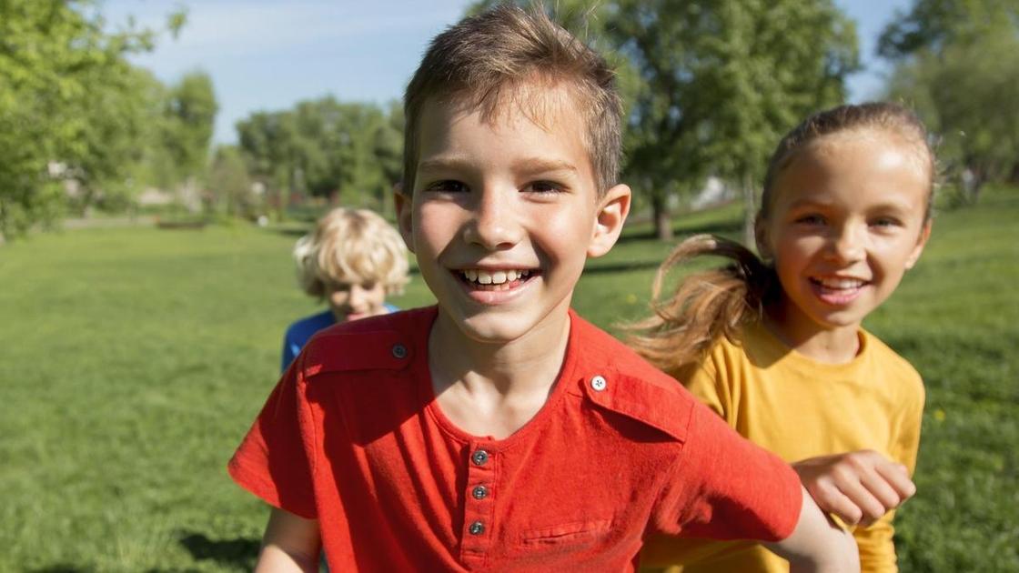 Мальчик в красной футболке бежит впереди и улыбается. Сзади его догоняет девочка в желтой футболке и мальчик в синей футболке