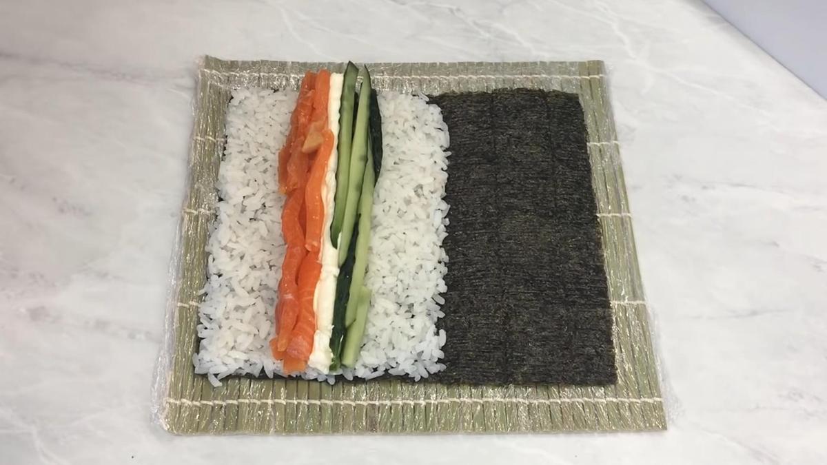 На листе нори с рисом выложены слабосоленый лосось, сливочный сыр, нарезанный огурец