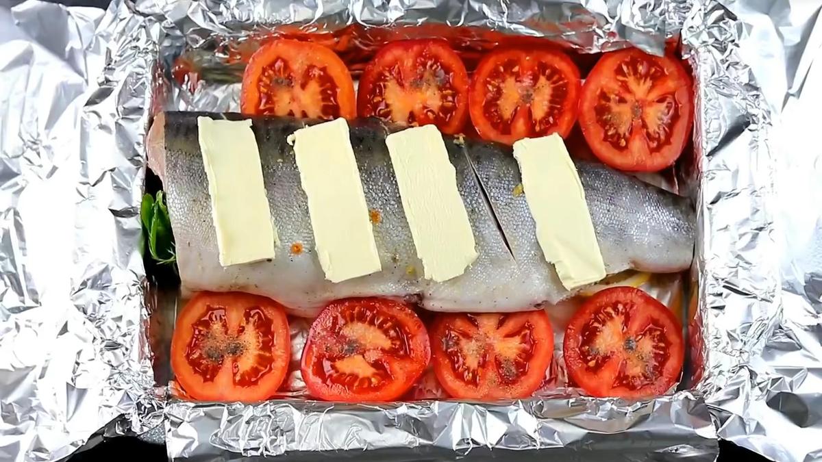 Рыба на противне, застеленном фольгой. Рядом помидоры. На рыбе сливочное масло