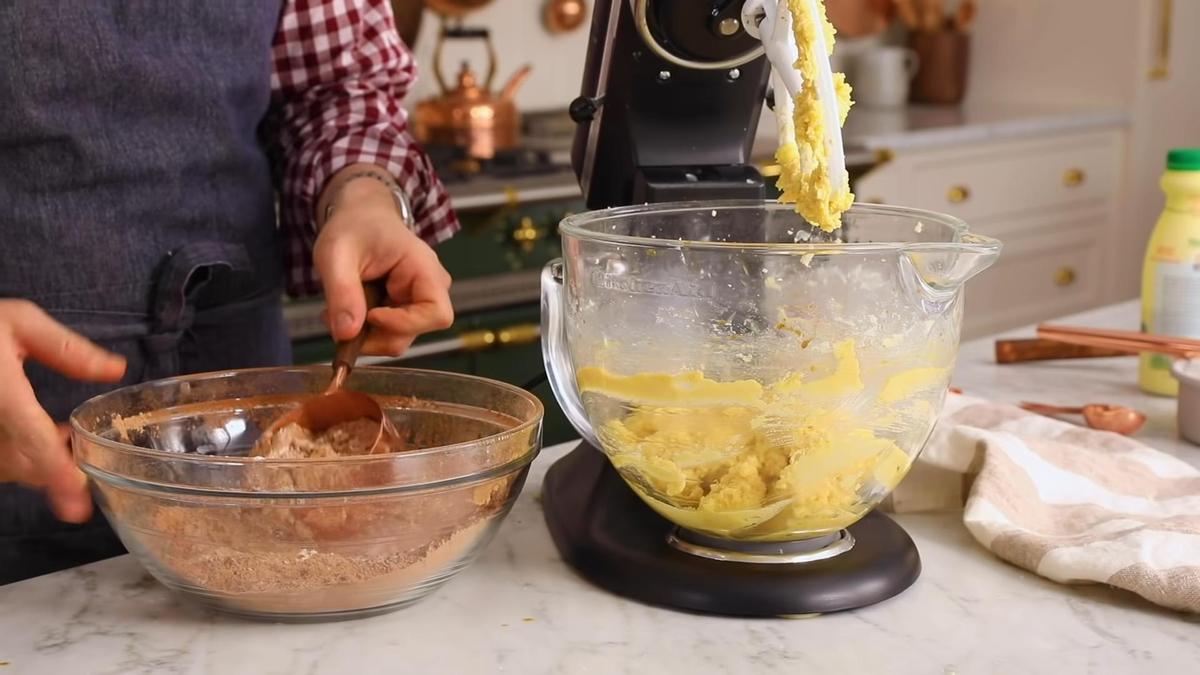 На столе миска со смесью сухих ингредиентов и миксер с чашей, в которой взбито масло