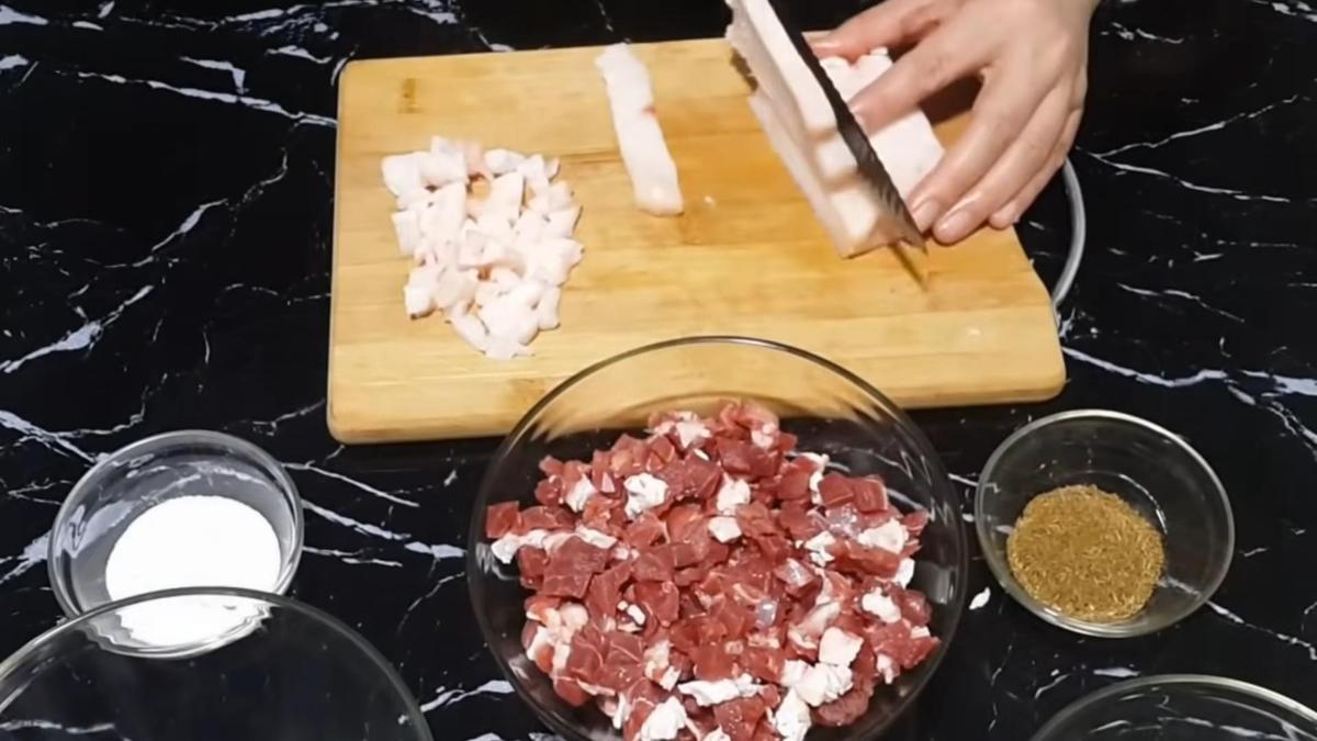 Курдючный жир нарезают кубиком на разделочной доске. Рядом тарелка с нарезанной бараниной
