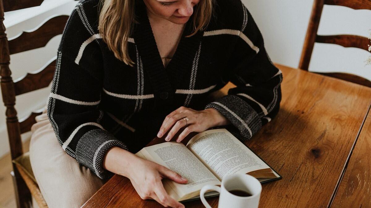 Девушка сидит за столом и читает книгу, на столе стоит чашка