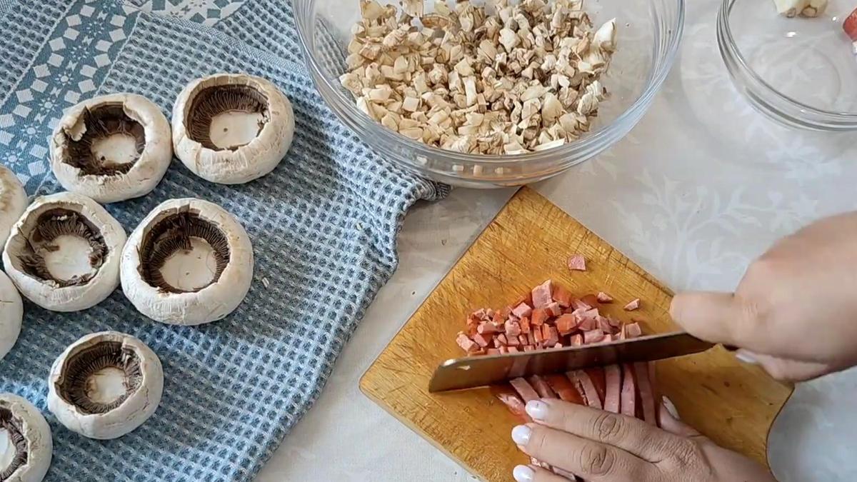 На разделочной доске нарезают сосиски. Рядом очищенные шляпки шампиньонов и нарезанные ножки грибов в миске