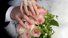 Руки молодоженов с обручальными кольцами скрещены и лежат на свадебном букете из розовых роз
