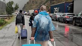 Машины стоят в пробке на границе, люди с чемоданами идут по дороге