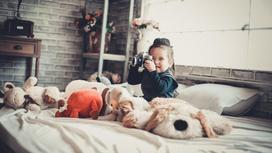 Девочка с фотоаппаратом сидит в кровати, окруженная игрушками