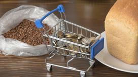 Маленькая корзина для продуктов наполнена монетами тенге и стоит рядом с хлебом и крупами