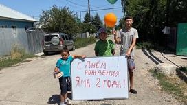 Дети стоят с плакатом, тортом и воздушными шарами возле ямы на дороге в Алматинской области и поздравляют ее с днем рождения