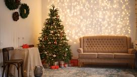 Новогодняя елка стоит на подставке в углу дома. Под стеной стоит диван, стул, столик, а на стене висят новогодние венки