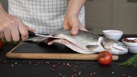 Рыбу разрезают ножом на дощечке. На столе рассыпаны горошины перца, лежит помидор, веточка укропа, стоят пиалы